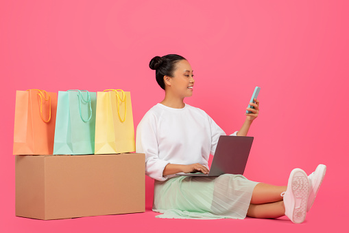 Asian woman sitting on floor among shopping bags, using laptop and smartphone, smiling korean female shopping online or managing small business, posing isolated on pink background in studio