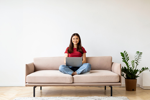 Happy young asian woman with laptop sitting cross-legged on sofa at home, smiling female in casual clothes working on computer in bright minimalist living room interior, copy space