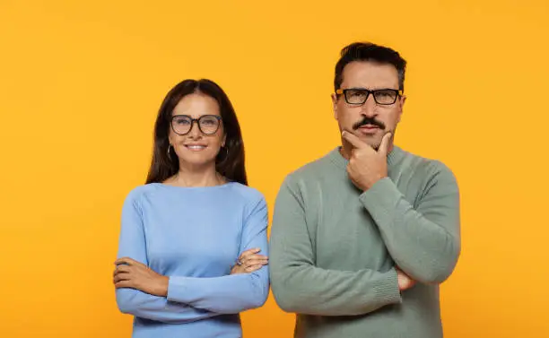 A contemplative caucasian man and a confident woman stand side by side, arms crossed, with the man touching his chin in thought and the woman smiling against an orange backdrop