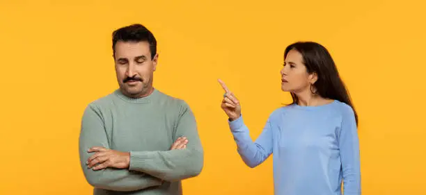 A caucasian man with a skeptical expression crosses his arms while a woman points a finger upwards, suggesting an argument or a discussion, against a bright orange background