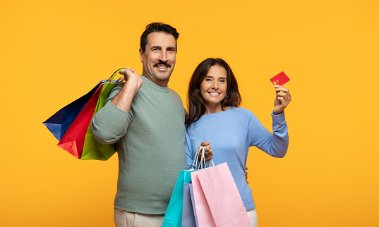A joyful glad old european couple with a man holding colorful shopping bags and a woman holding a red credit card, both smiling and posing against a vibrant yellow background
