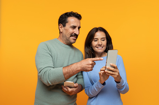 Positive old european family watch video on phone, point finger at smartphone, isolated on orange studio background. Relationships, sale app, ad and offer, fun and video call