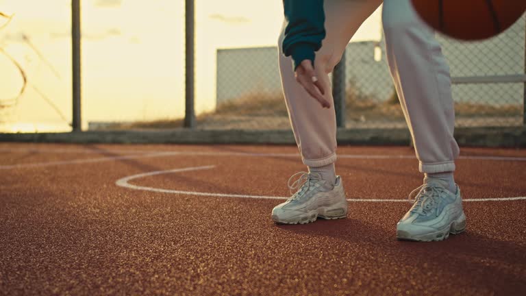 Close-up A girl in white sneakers and a sports uniform hits an orange ball from the floor on the Red street court for basketball players in the morning
