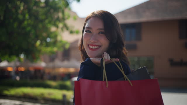 Happy young Asian woman holding shopping bag over her shoulder spinning around herself, stops to look at camera smiling