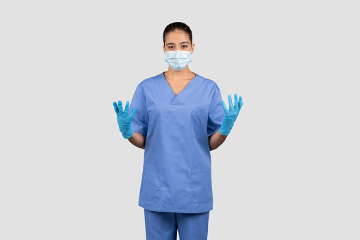 A composed european lady doctor in blue scrubs and protective gloves raises her hands, showcasing readiness for a sterile procedure or patient examination, isolated on gray studio background