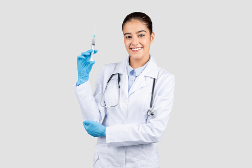 Positive young european doctor in a lab coat holding a syringe, showcasing readiness for a medical procedure with a friendly demeanor and professional expertise, isolated on gray studio background