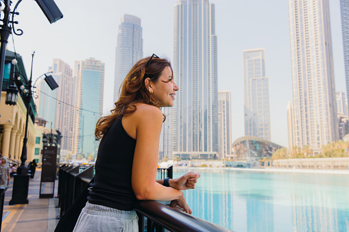 Side view of smiling west asian successful businesswoman looking at the central district of Dubai enjoying view of Skyscrapers by the waterfront during sunny day