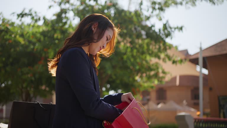 Happy young Asian woman in business casual clothes pulling out  her recently purchased clothes to check and admire while sitting on a bench at an outdoor shopping plaza on a bright sunny day