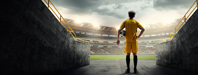 Young guy, soccer player in yellow uniform standing with football ball and looking at 3D stadium. Taking breath before game. Concept of live sport event, championship, match, game and tournament