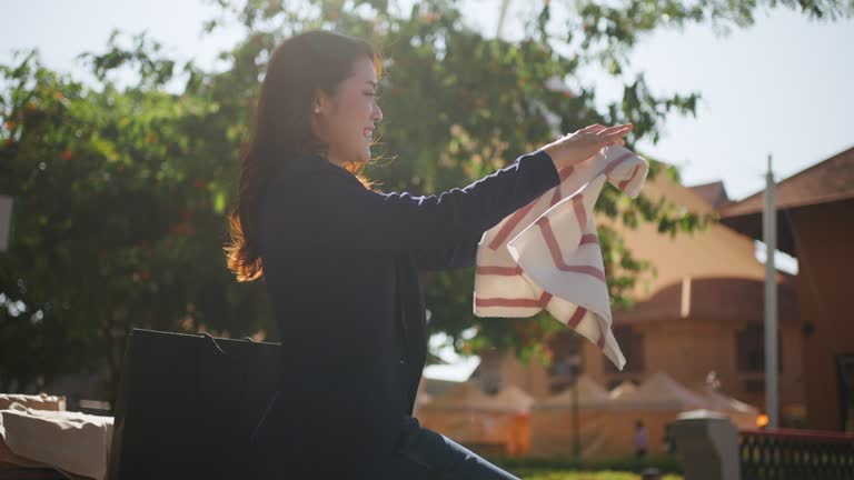 Happy young Asian woman in business casual clothes pulling out  her recently purchased clothes to check and admire while sitting on a bench at an outdoor shopping plaza on a bright sunny day