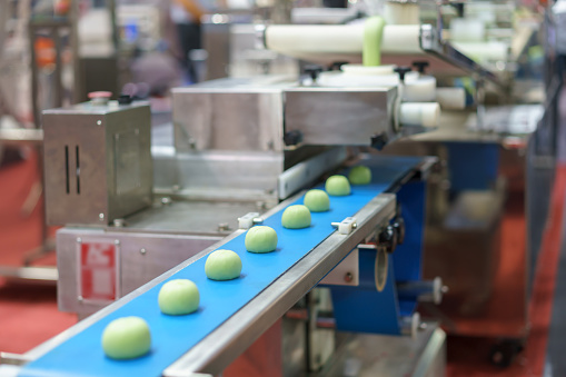 Close-up image of an automated industrial food processing system showing dough portions on a blue conveyor belt, with machinery elements in the background