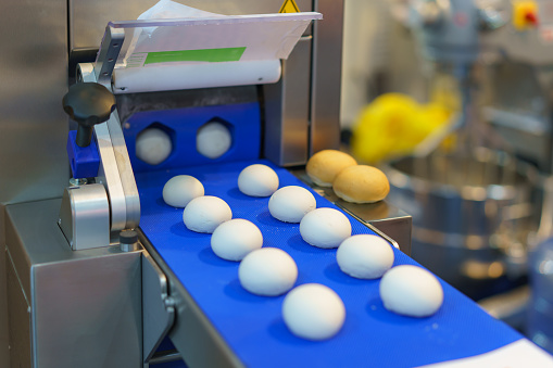 Close-up image of an automated industrial food processing system showing dough portions on a blue conveyor belt, with machinery elements in the background