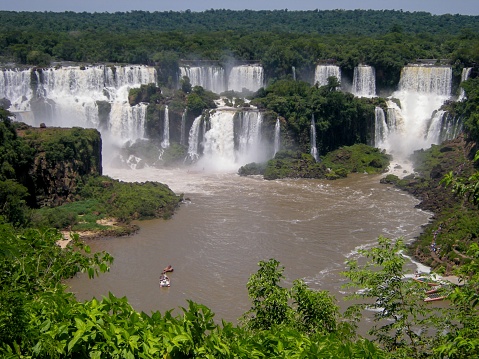 A young woman stands on a viewing platform on the Brazilian side of Iguazu Falls, smiling happily at the numerous waterfalls and lush landscape on the Argentinian side.