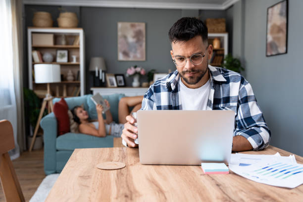 istock Young nervous concentrated man working on laptop calculating home finances while woman lying on sofa reading and surfing the net. Domestic life of freelancer worker and his girlfriend. 1997556204