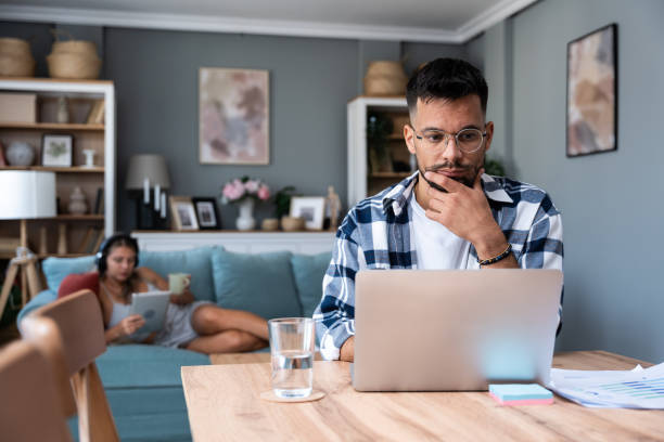 istock Young nervous concentrated man working on laptop calculating home finances while woman lying on sofa reading and surfing the net. Domestic life of freelancer worker and his girlfriend. 1997556254