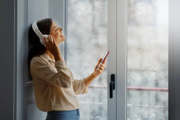 Young woman wearing wireless headphones listening music on smartphone at home, calm smiling female standing by rain-drenched window, showcasing a moment of relaxing domestic leisure, copy space
