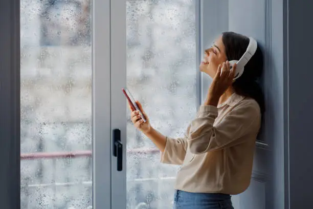 Smiling young woman with closed eyes listening music with stylish white headphones, beautiful female holding smartphone while standing beside a rain-streaked window, embodying relaxation