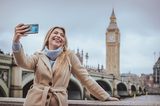 Candid portrait of a young cheerful blond woman in her 30s taking selfie with her smart mobile phone while laughing loud visiting down town city of London, England. Selective focus on the model with plenty of copy space on the background, which is defocused Westminster Bridge, Parliament building and Big Ben clock tower. Photo created during cold season outdoors and the model is with warm casual clothes on a cloudy day  - creative stock photo