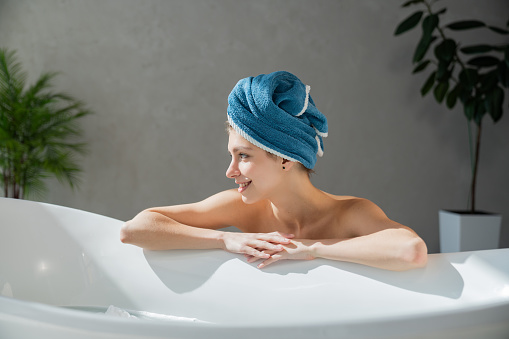 Happy female model with wrapped head, leaning on edge of bathtub in morning. Side view of dreamy caucasian girl looking to side, while getting ready to take bath at home. Spa, daily routine concept.