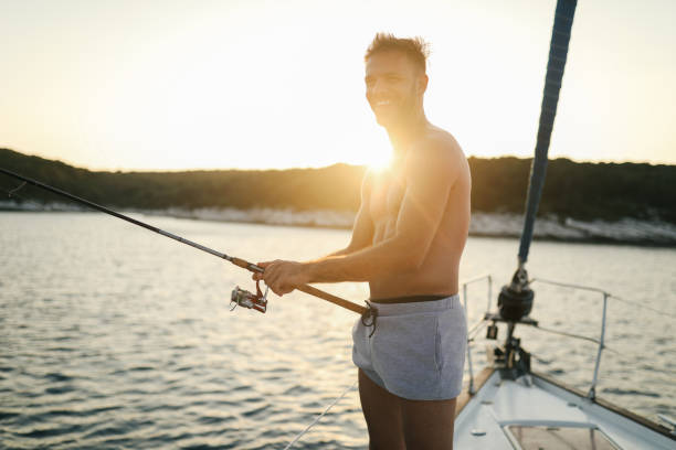 Young handsome man fishing from a sailboat deck, shirtless, he is smiling, looking happy and excited.