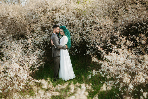 a bearded groom plays and a girl with green hair against the background of blooming spring white bushes