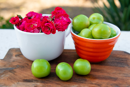 Front view of three, green plums laying on wet, wooden board, bowls with pink roses and green plums behind. Concept of summer.
