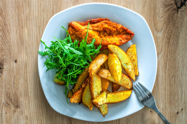 Plate with baked potatoes, meat, green fresh arugula leaves on dining table. Dish with hot oven roasted potato wedge, chicken breast, rucolla, spices on wooden table. Gourmet baking dinner, top view