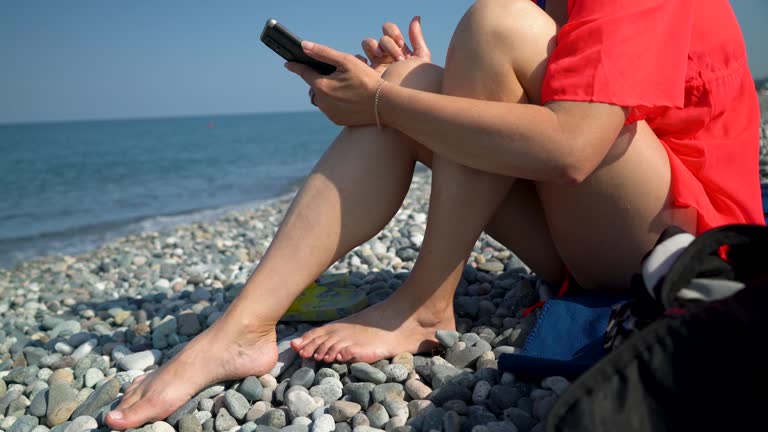 An attractive woman sits on the seashore and looks at her smartphone.