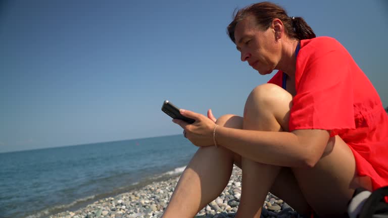 An attractive woman sits on the seashore and looks at her smartphone.