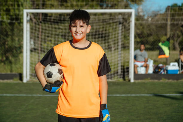 Confident young boy in goalkeeper outfit holds soccer ball on sunlit field with net background.