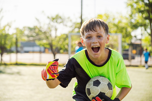 Excited boy in goalkeeper gear holding a soccer ball on a field, expressing triumph and happiness.