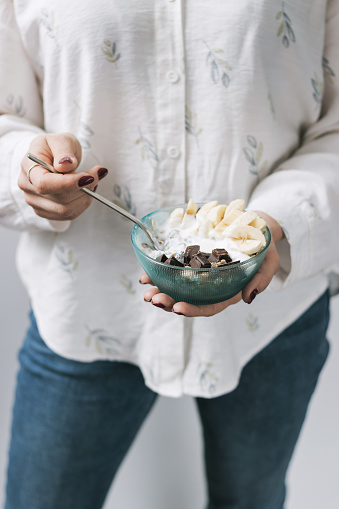 Woman eating healthy food for breakfast, cereal, granola, muesli, oatmeal with banana, dark chocolate and yogurt in a bowl.