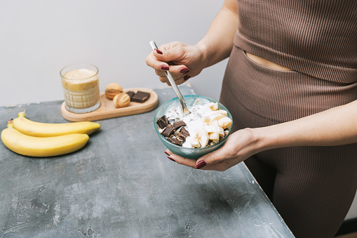 Athletic woman in sportswear eating healthy food for breakfast, cereal, granola, muesli, oatmeal with banana, dark chocolate and yogurt in a bowl.