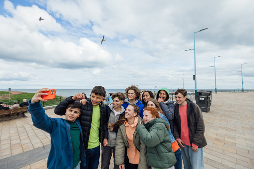 A group of male and female teenagers enjoying a weekend hanging out together in Whitley Bay, North East England. They are standing outside with the coastline behind them while they take a selfie on a mobile phone.