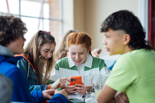 A group of male and female teenagers enjoying a weekend hanging out together in Whitley Bay, North East England. They are sitting in a cafe, enjoying food and drink while two girls are looking at a mobile phone together and smiling.