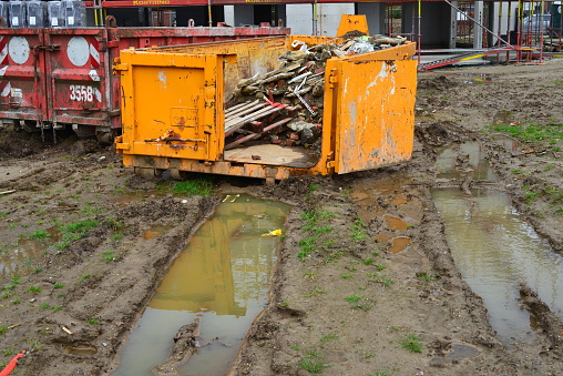 Leuven, Flemish-Brabant, Belgium - February 9, 2024: mud earth from rain in front of a yellow metal industrial container filled with garbage from construction site apartment blocks