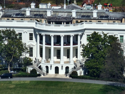 This image shows the White House, the official residence and workplace of the President of the United States, viewed from an angle that highlights the South Portico. The South Portico features a prominent semi-circular portico with tall, white columns and a set of stairs leading to a balcony above the entrance.  The building itself is grand with its neoclassical architecture, boasting numerous windows and intricate details. The white paint of the exterior contrasts with the green of the well-manicured lawn in the foreground. On the roof, there are several mechanical units and what appear to be maintenance workers present. There are a couple of vehicles near the entrance and some foliage that partially obscures the view on the left-hand side of the image. The sky above is clear, indicating fair weather and adding a serene backdrop to this iconic structure.