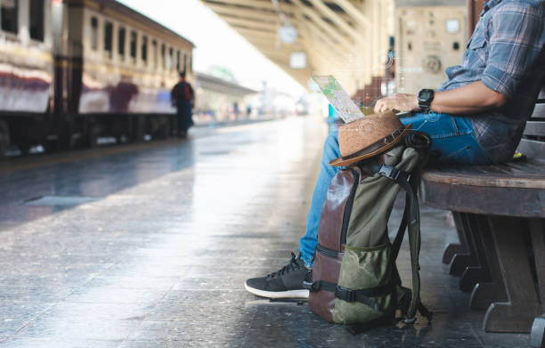 Digital normad waits for a train while seated on a bench at a train station, typing on a laptop and looking up a location on a map with a rucksack.