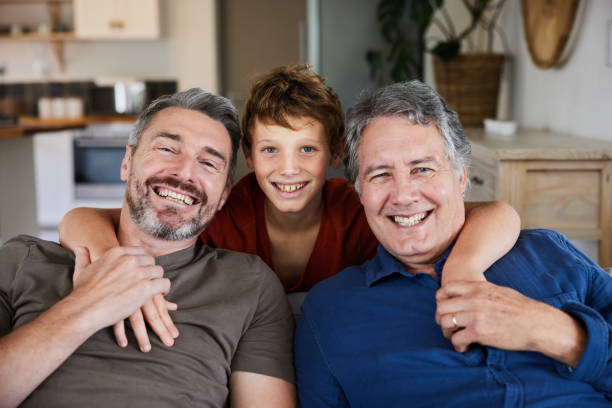 Smiling man sitting with his young son and mature dad on a sofa Portrait of a cute young boy smiling with his father and grandfather in their living room at home during a visit father son grandfather fathers day stock pictures, royalty-free photos & images