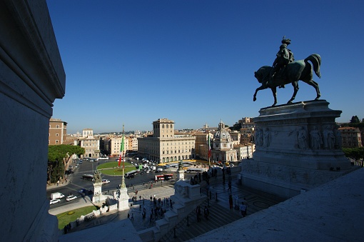 The picture portrays a sweeping, elevated view of a historic cityscape under a bright blue sky. On the right side is a dark bronze equestrian statue, capturing a mounted figure in silhouette against the sky. Below, a wide set of stairs descends towards a bustling square filled with people, traffic, and classic architecture. The square acts as a hub, with roads radiating out from it, flanked by majestic buildings and monuments. The city stretches into the distance, suggesting an expansive and culturally rich urban environment.