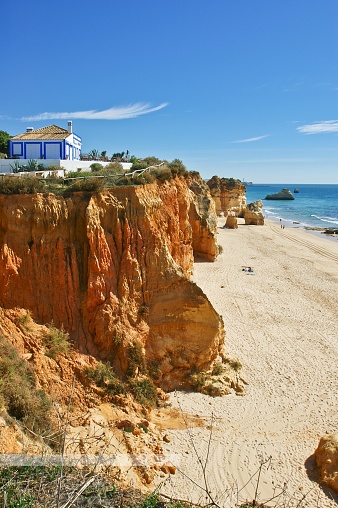 The picture shows a stunning beachscape. On the right side, towering, rugged orange cliffs dominate the foreground, leading down to a golden sandy beach below. The cliffs are jagged and steep, showcasing the natural erosion from the sea. In the distance, there are some large rocks scattered in the turquoise ocean waters, which gently lap against the shore.  To the left, atop the cliff, there's a white house with blue trim, typical of Mediterranean or coastal architecture. The house stands out against the natural colors of the surrounding landscape. Vegetation can be seen sporadically along the cliff edges and on the house's property, adding a touch of green to the scene.  The sky is a clear, azure blue, indicating good weather, and there is ample sunlight casting shadows on the cliff face, enhancing the textures and contours of its surface. On the beach, a few people are visible, suggesting that the area is somewhat serene and not overly crowded. The overall scene invokes a sense of peace and natural beauty, inviting the viewer to imagine the sound of the waves and the warmth of the sun.