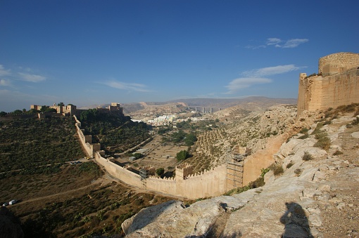 This picture appears to capture a historical fortress situated on a hilltop. The fortress is characterized by tall, sand-colored walls and a series of towers. A fortified wall snakes down the hill on the left side, suggesting that the defense system was extensive and well-planned to protect the complex and surrounding area.  The photo is taken during the day under clear, blue skies. The natural landscape surrounding the fortification is arid and features sparse vegetation, rolling hills, and what appear to be rocky outcrops. In the distance, modern structures can be seen, including what looks like towers and buildings, indicating the presence of a contemporary town or city.  Shadowing in the foreground suggests the sun is high and might be casting the photographer's shadow onto the rocky ground, indicating the photo was likely taken in the afternoon. Overall, the image portrays a mix of ancient architecture and modern development, encapsulating a place where historic significance coexists with present-day life.