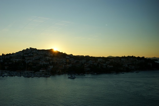 This picture shows a serene sunset view over a coastal town. The sun appears to be just at the cusp of the hill, creating a silhouette of the landscape. At the top of the hill, you can see clusters of buildings, possibly residential homes, neatly arranged and possibly bathed in the fading sunlight. In the foreground, a calm body of water - likely a bay or harbor - reflects the waning light, and several boats or yachts are anchored, hinting at a life of maritime leisure. There's a clear sky with minimal cloud cover, and the overall mood is peaceful and reflective.