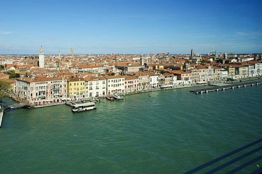 This is an image of the Grand Canal in Venice, Italy, taken from an elevated vantage point during the daytime. The canal winds through the city and is flanked on either side by rows of multi-storied buildings exhibiting the classic Venetian architecture with colorful facades, ornate windows, and terraces. The buildings are tightly packed, creating a continuous urban scene along the water's edge. Several boat-shaped yellow vessel, which appear to be public waterbuses (vaporetti), are docked at designated stops or cruising along the canal.  The water of the canal has a greenish-blue hue, reflecting the clear sky above. Across the canal, we can see several prominent buildings and bell towers rising above the uniformly high rooftops. One notable tower on the right of the image stands taller than its surroundings, which could be a campanile, a typical feature in many Italian cityscapes. In the far distance, there is a clear view of the horizon where the sky meets the outline of the city's structures. A few sparse mooring poles are spaced out in a line parallel to the buildings' facade on the water.  The weather appears pleasant with a clear, bright blue sky with a few wispy clouds, indicating it's likely a sunny day, which contributes to the vividness of the colors in the photograph.