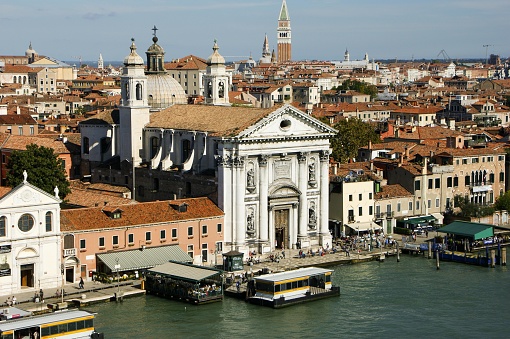 The picture depicts a grandiose, white facade of a classic European-style building with intricate architectural details, including statues and columns. This building appears to be a church or a cathedral due to its style and the presence of a religious cross at its peak. It stands prominently at the edge of a waterway, suggesting it could be in a coastal or canal-based city like Venice in Italy.   In the background, numerous other red-tiled roof buildings spread out, varying in height and size, suggesting a dense, historic urban area. The image also shows a busy waterfront with several boats and a ferry station, indicating this is likely a popular area for both local transportation and tourism. The calm water reflects the beauty of the buildings and the variety of activities along its edge. The sky is a clear blue and suggests a sunny, pleasant day.