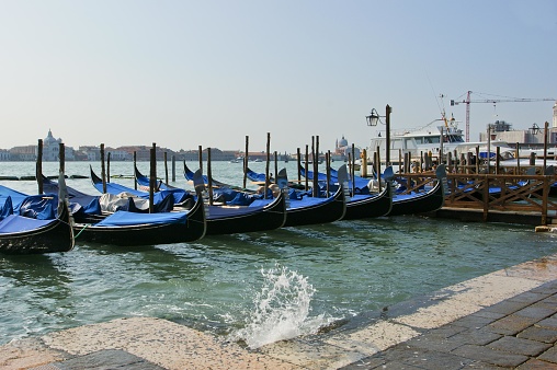 This picture shows a row of gondolas moored alongside a wooden dock on the edge of a body of water. The gondolas have blue covers on them and are characteristic of those found in Venice, Italy. In the background, there is a construction site with a crane, indicating some kind of development or maintenance work is being carried out. On the left side of the image, there is a splash of water, suggesting movement or activity in the water. The sky is clear with an appearance of good weather. The overall scene is calm with no visible people, focusing on the iconic gondolas lined up along the waterfront.