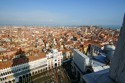 This is an aerial photograph of Venice, Italy. The image provides a sweeping view over the historic city, with its dense arrangement of buildings, narrow alleys, and canals. On the left, you can see the famous St. Mark's Square (Piazza San Marco) with the recognizable patterned paving, crowded with people. Adjacent to the square is the St. Mark's Basilica with its easily identifiable Byzantine domes. To the right is the Doge's Palace (Palazzo Ducale) with its Gothic architectural features. In the distance, the Venetian Lagoon glistens under the bright blue sky. The city's red rooftops and the campaniles (bell towers) rising from the sea of buildings add to the characteristic Venetian skyline. The photo is taken during the day under clear weather conditions, providing a clear view of the city sprawled out to the horizon.