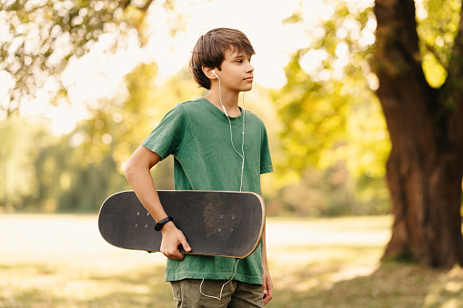 Handsome thoughtful teen boy ia walking while holding his skateboard in a green park.