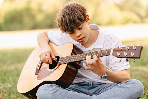 Handsome teen boy is learning how to play guitar while sitting in park on grass.