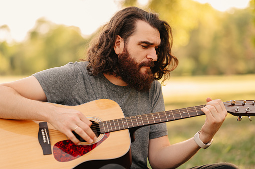 Close up shot of a handsome bearded man playing guitar while sitting in park in summer.